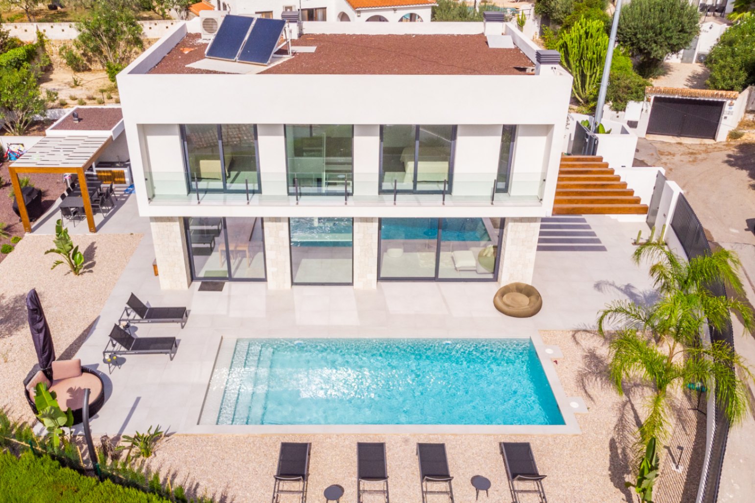 Vista aérea de la villa con piscina rectangular, rodeada de tumbonas y gravilla clara. Diseño moderno con paneles solares.