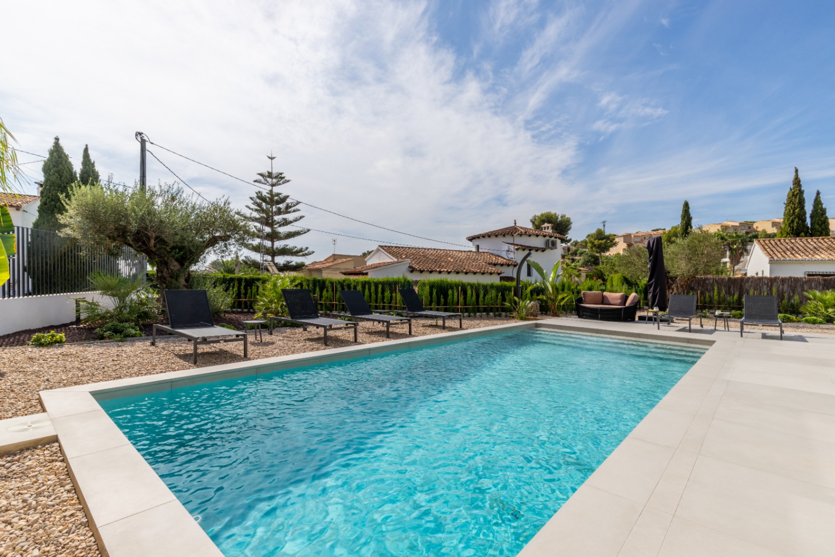 Vista de la piscina privada con borde de piedra clara, rodeada de tumbonas negras y jardines mediterráneos.