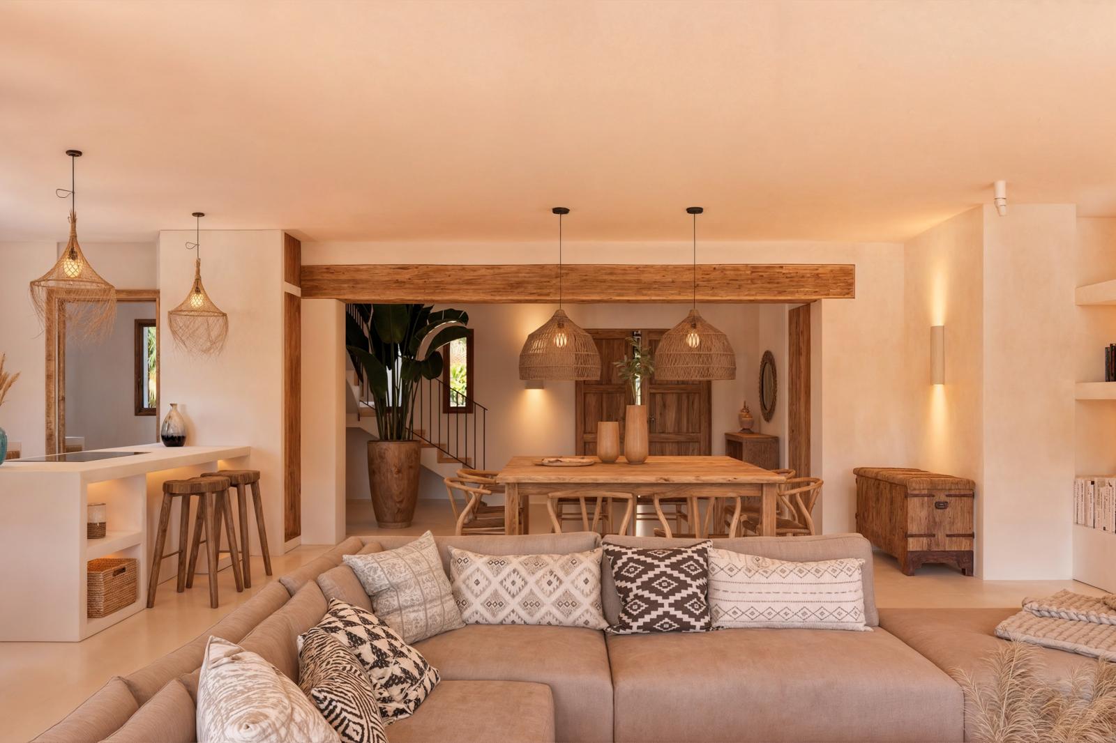 Salón-comedor de planta abierta con vigas de madera natural, lámparas de fibras vegetales y mobiliario de estilo rústico mediterráneo en tonos cálidos.