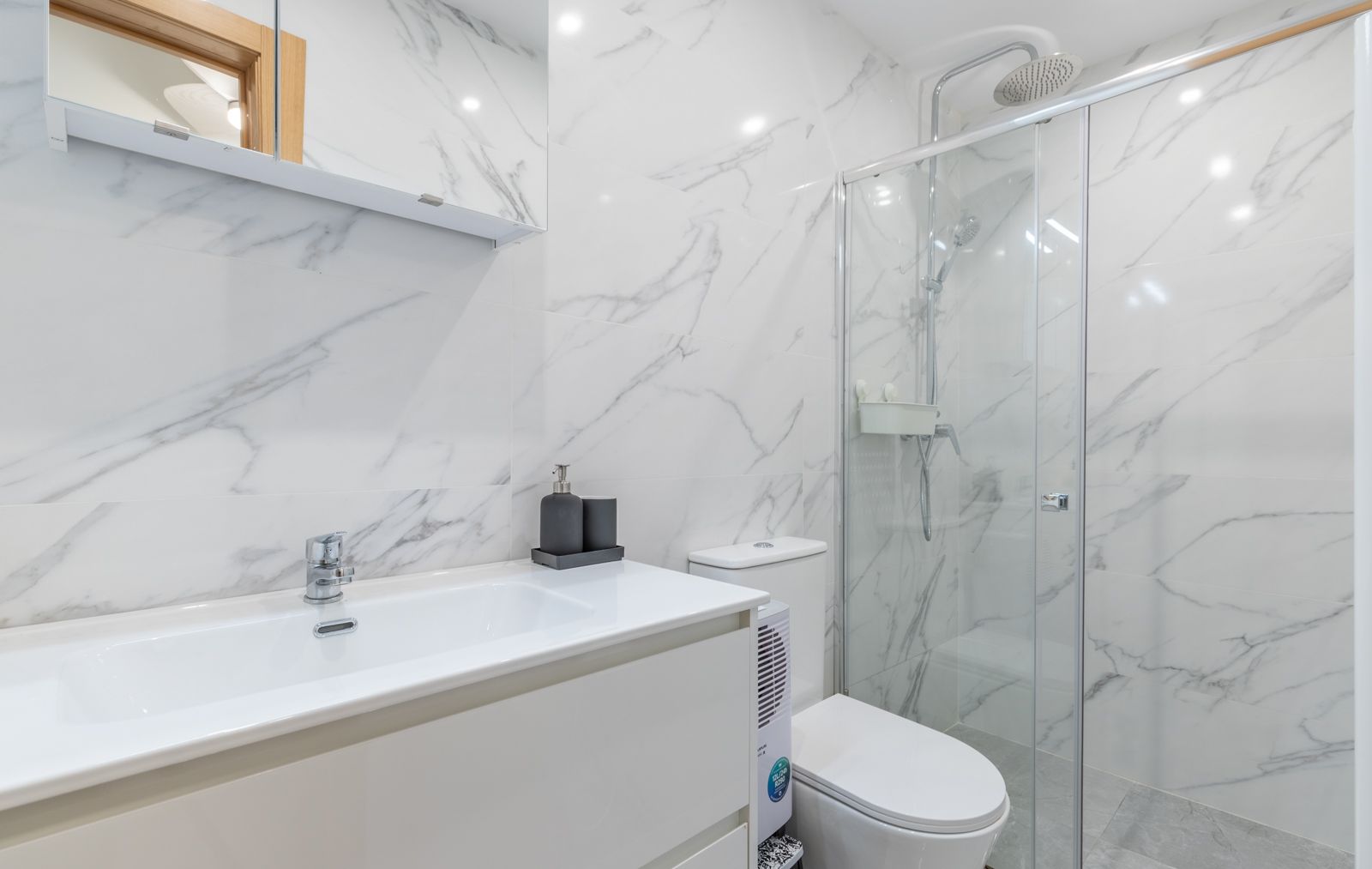 Baño contemporáneo con revestimiento de porcelánico efecto mármol, lavabo integrado en mueble blanco y ducha de obra con mampara de cristal y grifería cromada.