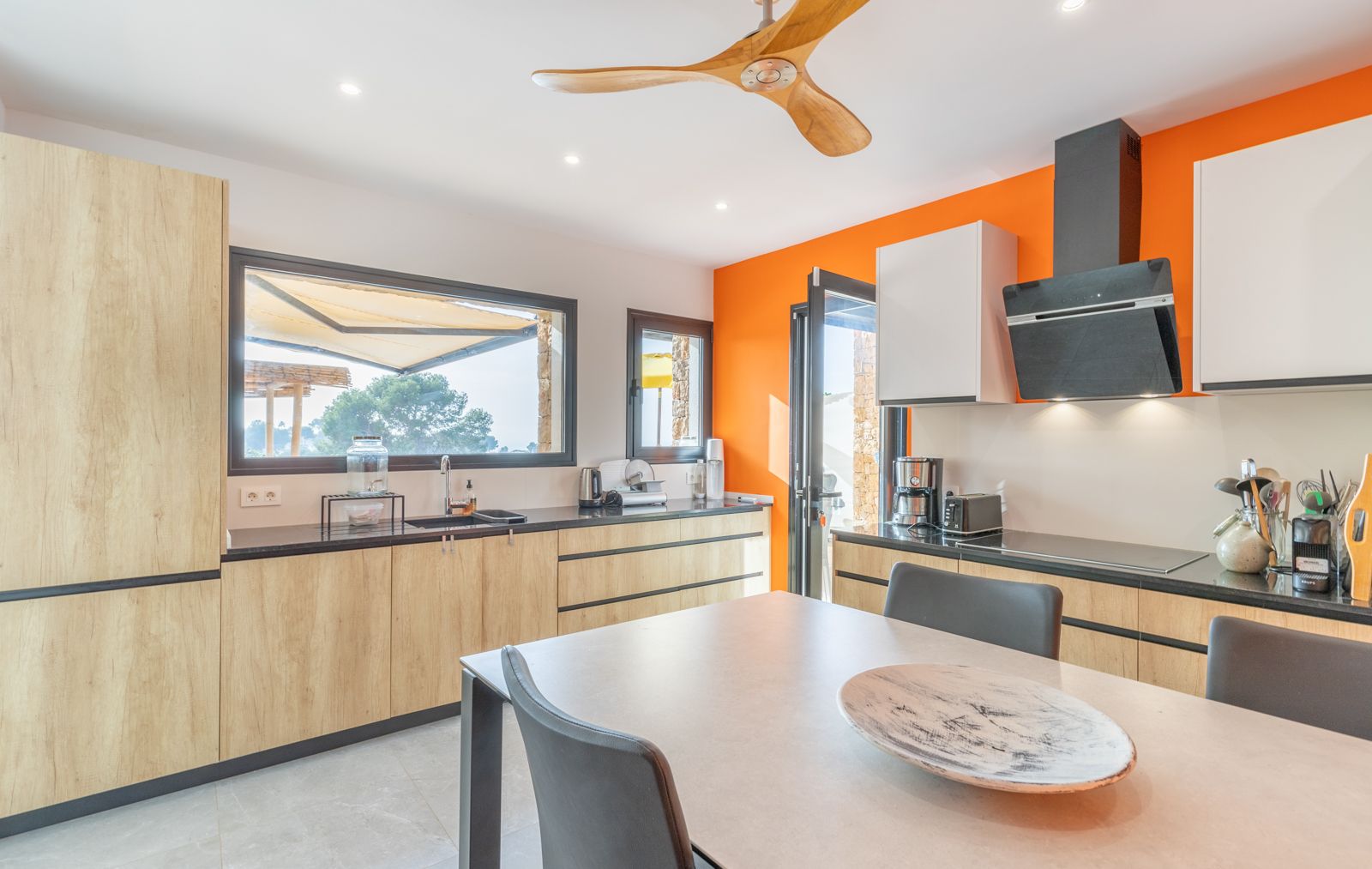 Cocina de diseño contemporáneo con mobiliario de madera clara, encimera oscura y pared de acento naranja. Dispone de ventilador de techo de madera y acceso directo a la terraza.