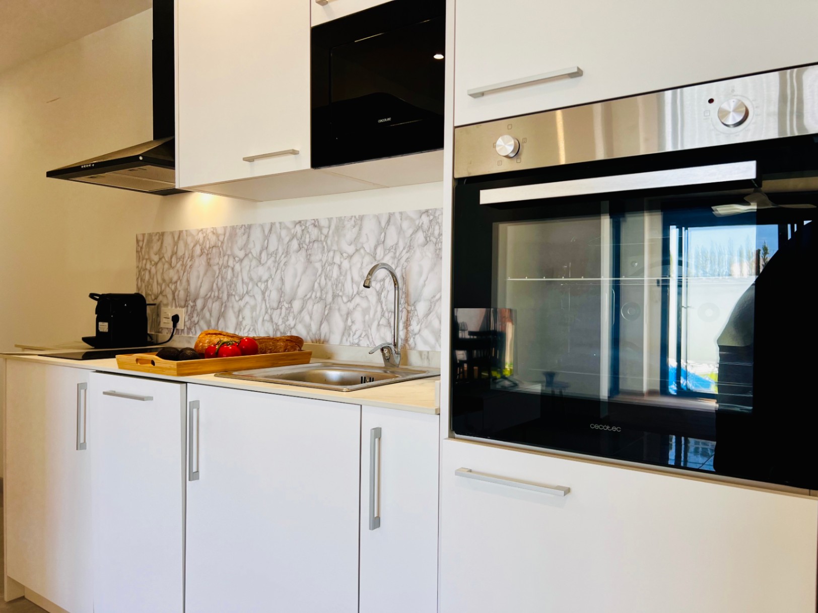 Cocina moderna con armarios blancos, electrodomésticos Cecotec integrados y un elegante salpicadero con efecto mármol. Incluye fregadero de acero inoxidable y campana extractora negra.