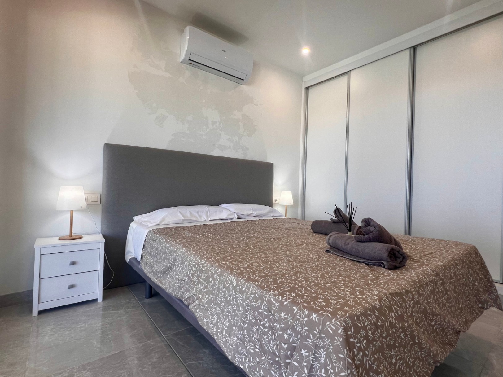 Dormitorio contemporáneo con armario empotrado de puertas correderas blancas, aire acondicionado y cabecero tapizado gris sobre suelo de gres.
