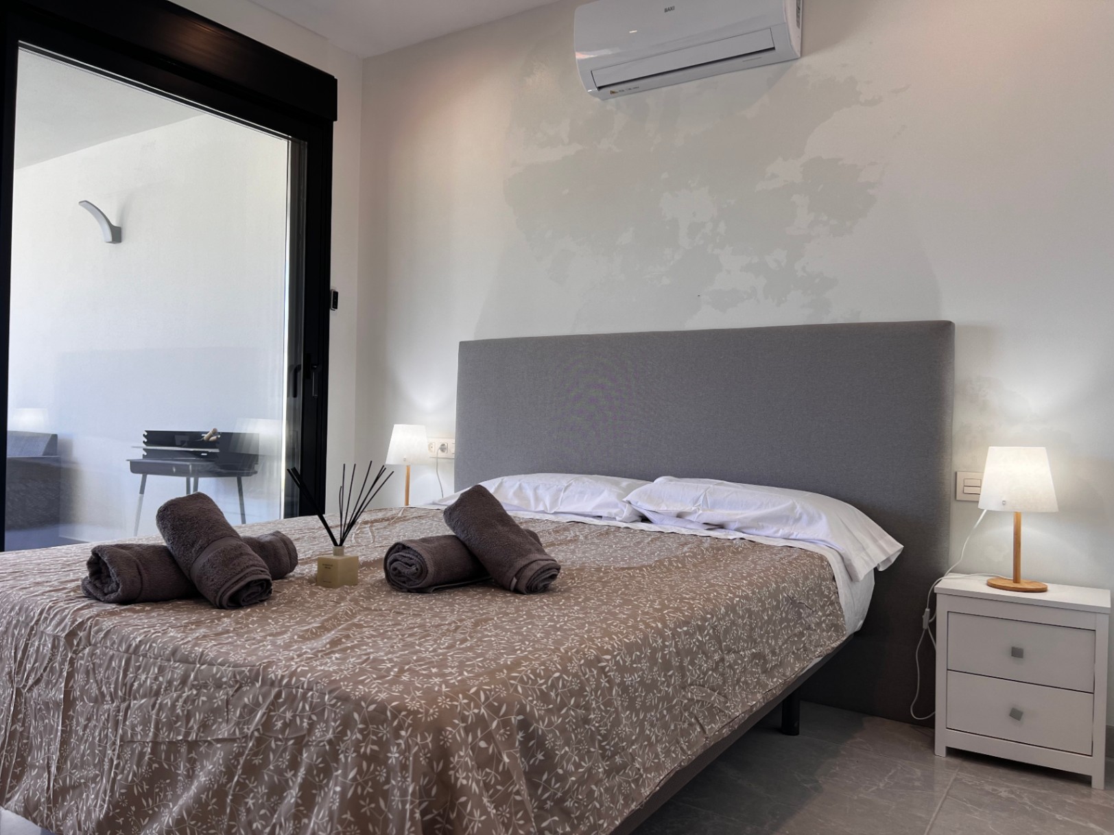 Dormitorio contemporáneo con cabecero textil gris, aire acondicionado tipo split y acceso directo a la terraza mediante grandes ventanales de marco negro.