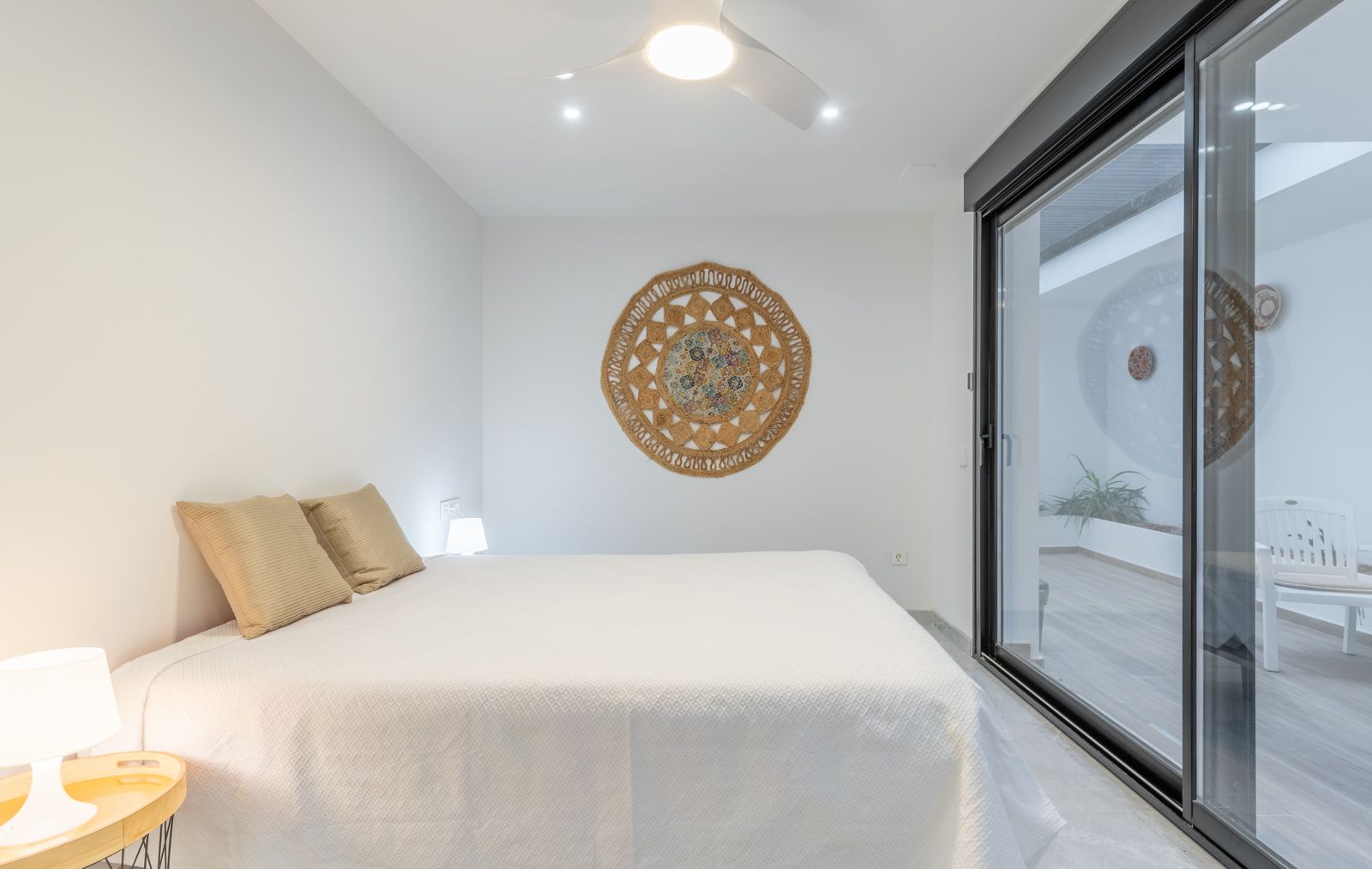 Dormitorio minimalista con acceso directo a la terraza mediante ventanales correderos, ventilador de techo con luz y decoración en fibras naturales sobre paredes blancas.