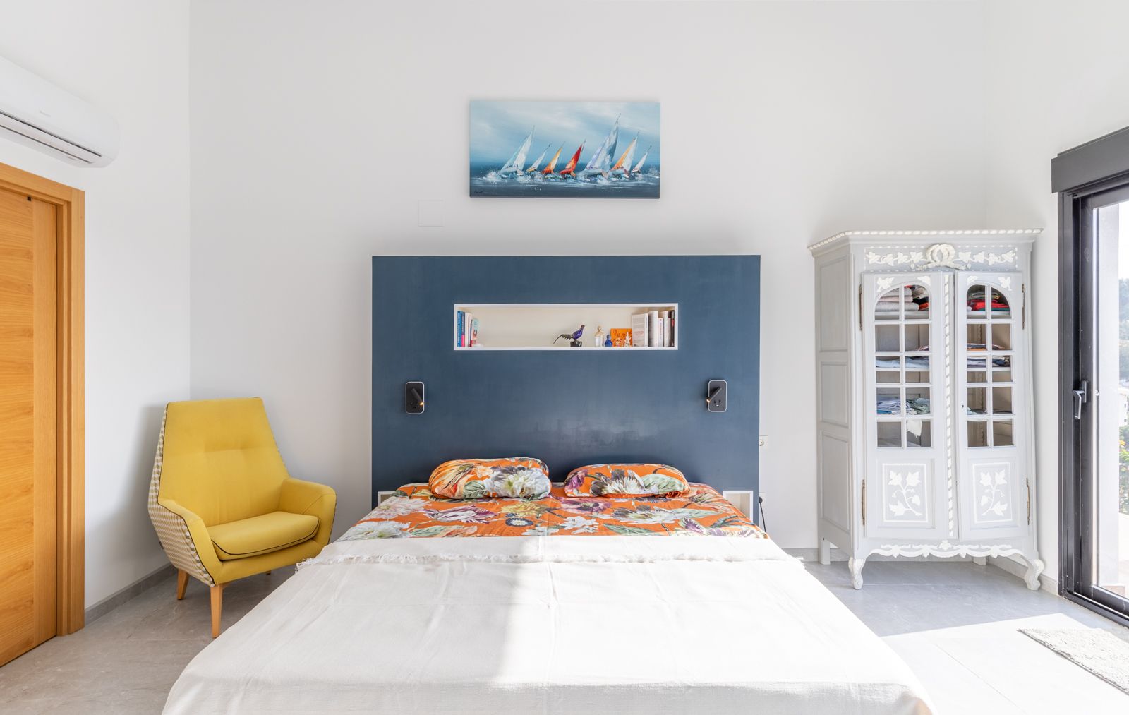Dormitorio moderno con cabecero azul integrado, sillón amarillo de diseño y armario clásico. Incluye aire acondicionado y acceso directo al exterior mediante ventanal.