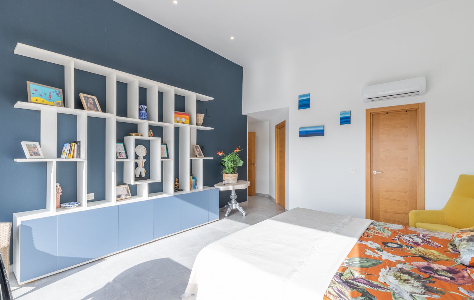 Dormitorio moderno con una estantería geométrica blanca sobre pared azul marino, armarios bajos integrados y suelos de gres porcelánico claro.