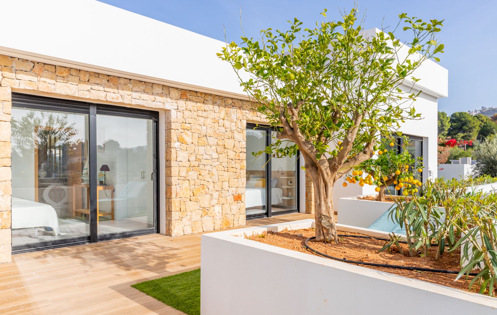 Fachada moderna con muros de piedra natural, ventanales de suelo a techo y jardineras integradas con un limonero, conectando los dormitorios con la terraza exterior.