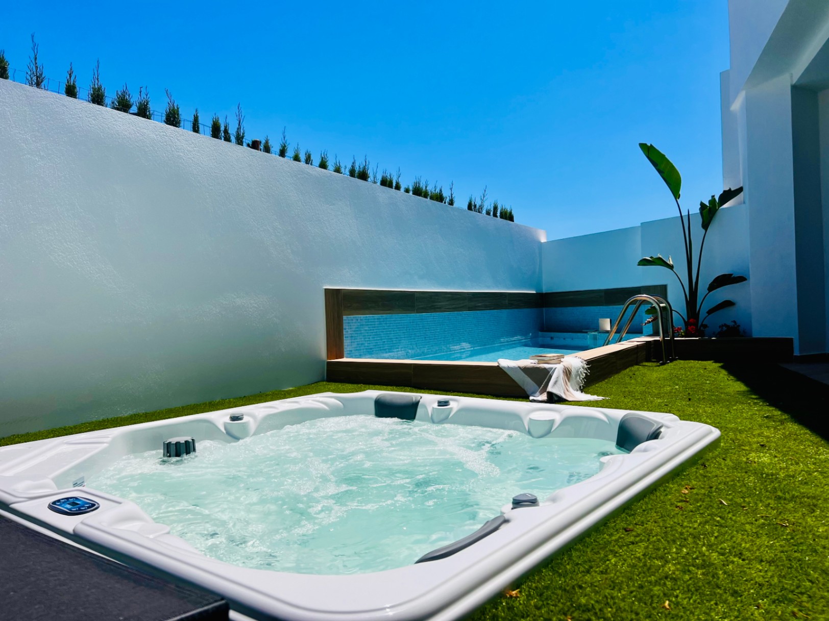 Jacuzzi exterior integrado en césped artificial, junto a una piscina privada con gresite azul y muros perimetrales blancos que garantizan privacidad.