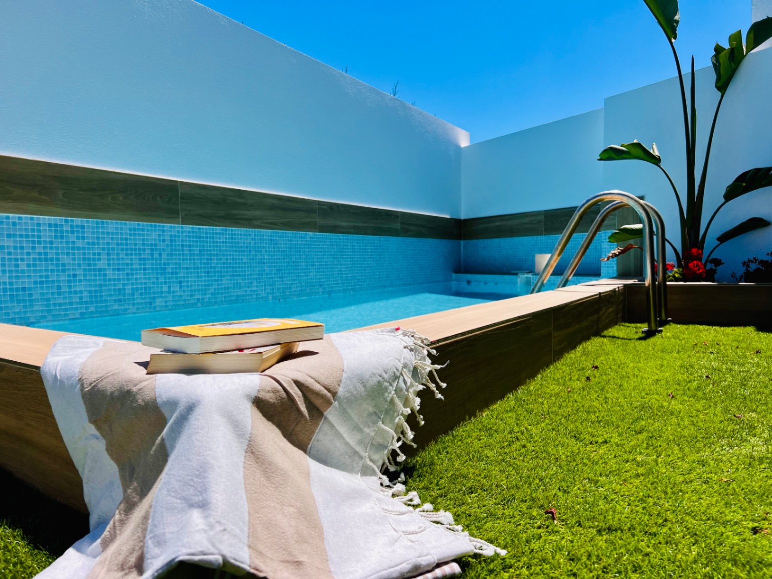 Jacuzzi privado con revestimiento de gresite azul, borde de madera y zona de césped artificial, rodeado de muros blancos bajo un cielo despejado.