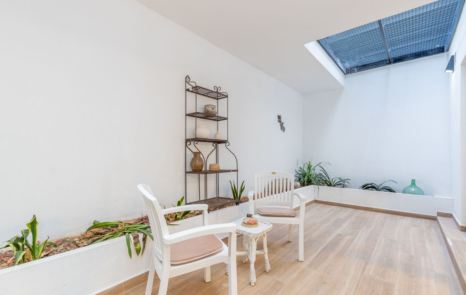Patio interior luminoso con claraboya, suelos de gres efecto madera, jardineras integradas y zona de descanso con mobiliario blanco y estantería de forja.