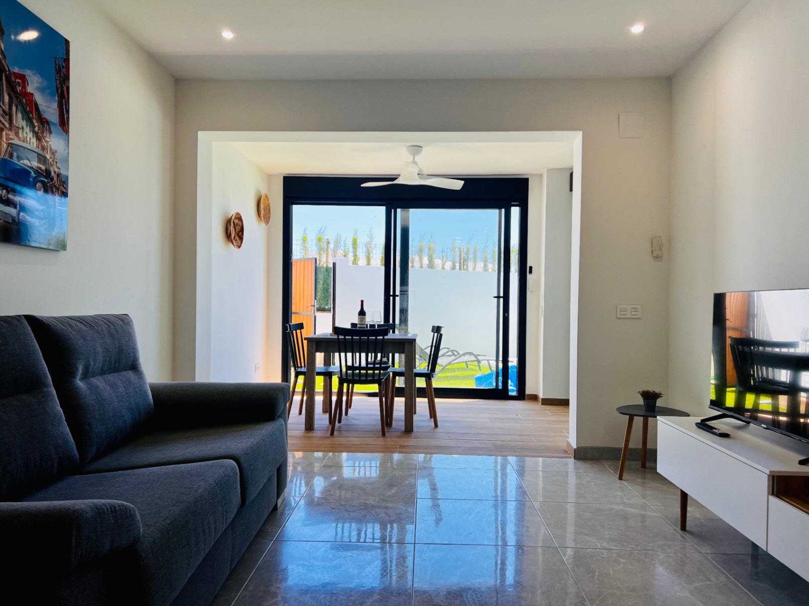 Salón contemporáneo con suelos de gres pulido, sofá en tonos grises y zona de comedor integrada con ventilador de techo y acceso directo al patio exterior.