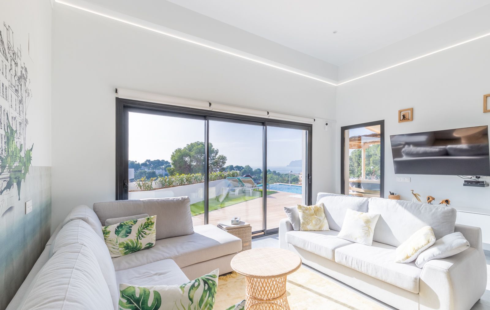 Salón luminoso con sofás grises, iluminación LED integrada y amplios ventanales que ofrecen vistas directas a la piscina, el jardín y el mar Mediterráneo.