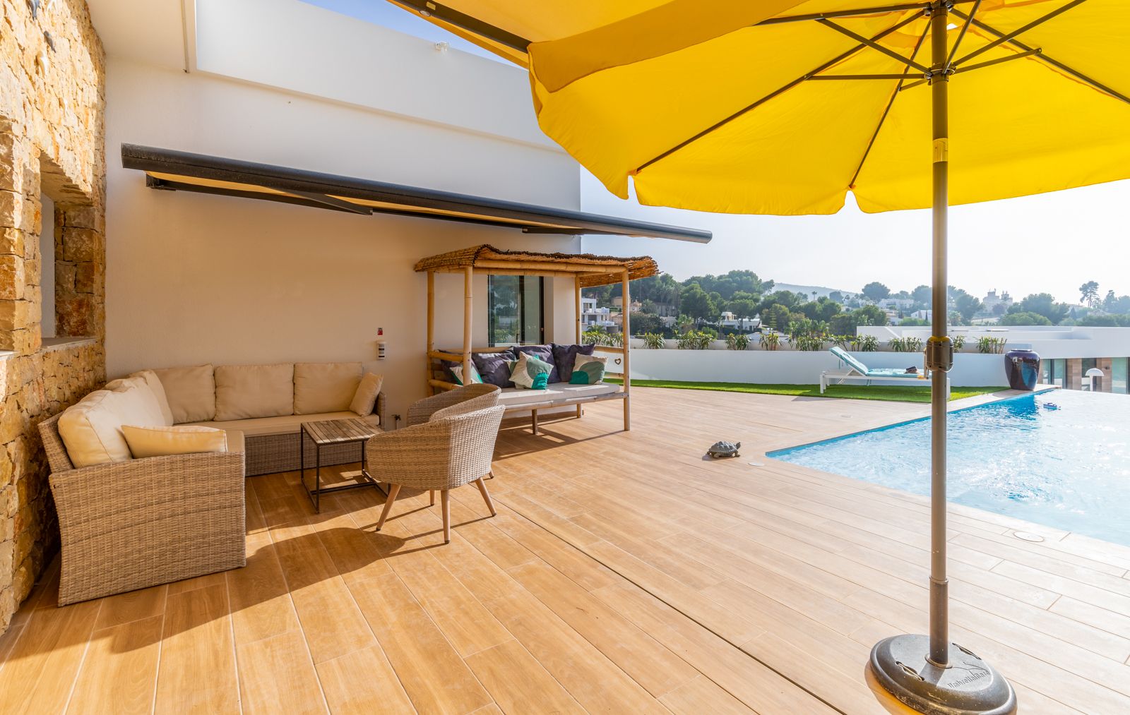 Terraza exterior con suelo de gres porcelánico imitación madera, zona de estar con sofás de mimbre, cama balinesa de bambú y piscina junto a un muro de piedra natural.