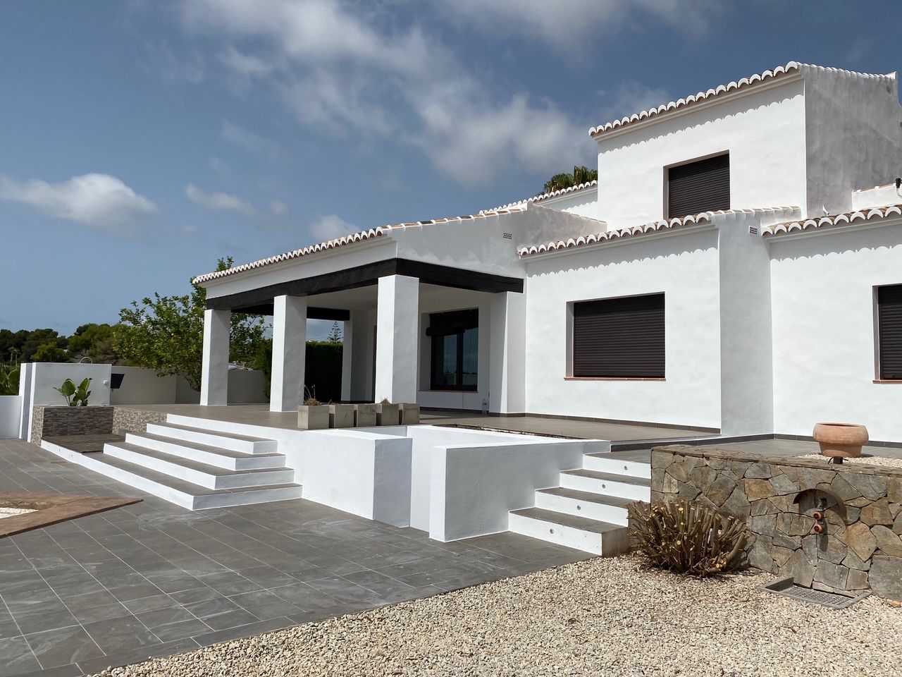 Fachada mediterránea blanca con vigas de madera oscura, amplia terraza de gres y muros de piedra natural. Incluye una zona de ducha exterior y escaleras de acceso integradas.