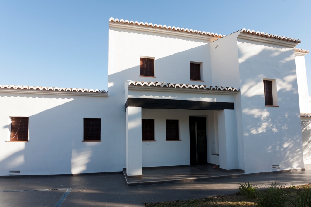 Fachada mediterránea de paredes blancas con carpintería de aluminio imitación madera, vigas oscuras y cubiertas de teja tradicional sobre un porche sombreado.