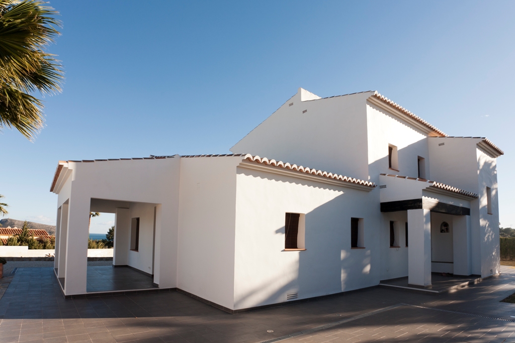 Vista exterior de la villa mediterránea con fachada blanca, tejados de teja y amplias terrazas pavimentadas que conectan con el porche cubierto y el entorno natural.