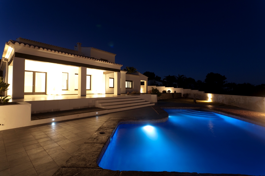 Vista nocturna de la fachada blanca iluminada y la piscina de forma irregular con iluminación subacuática azul, rodeada de una amplia terraza de gres.