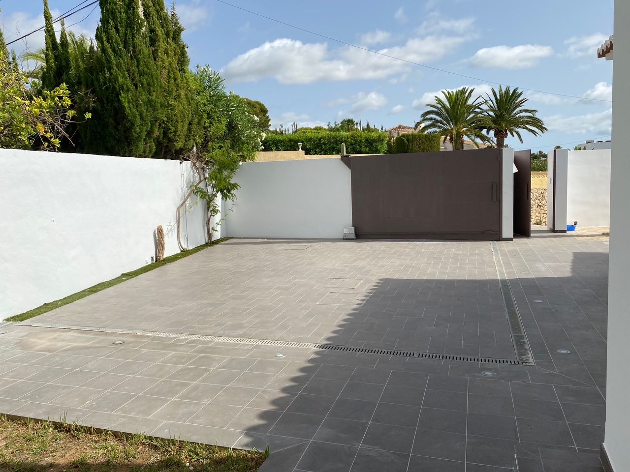 Zona de aparcamiento privada con suelo cerámico gris, puerta corredera motorizada y acceso peatonal, rodeada de muros blancos y vegetación mediterránea.