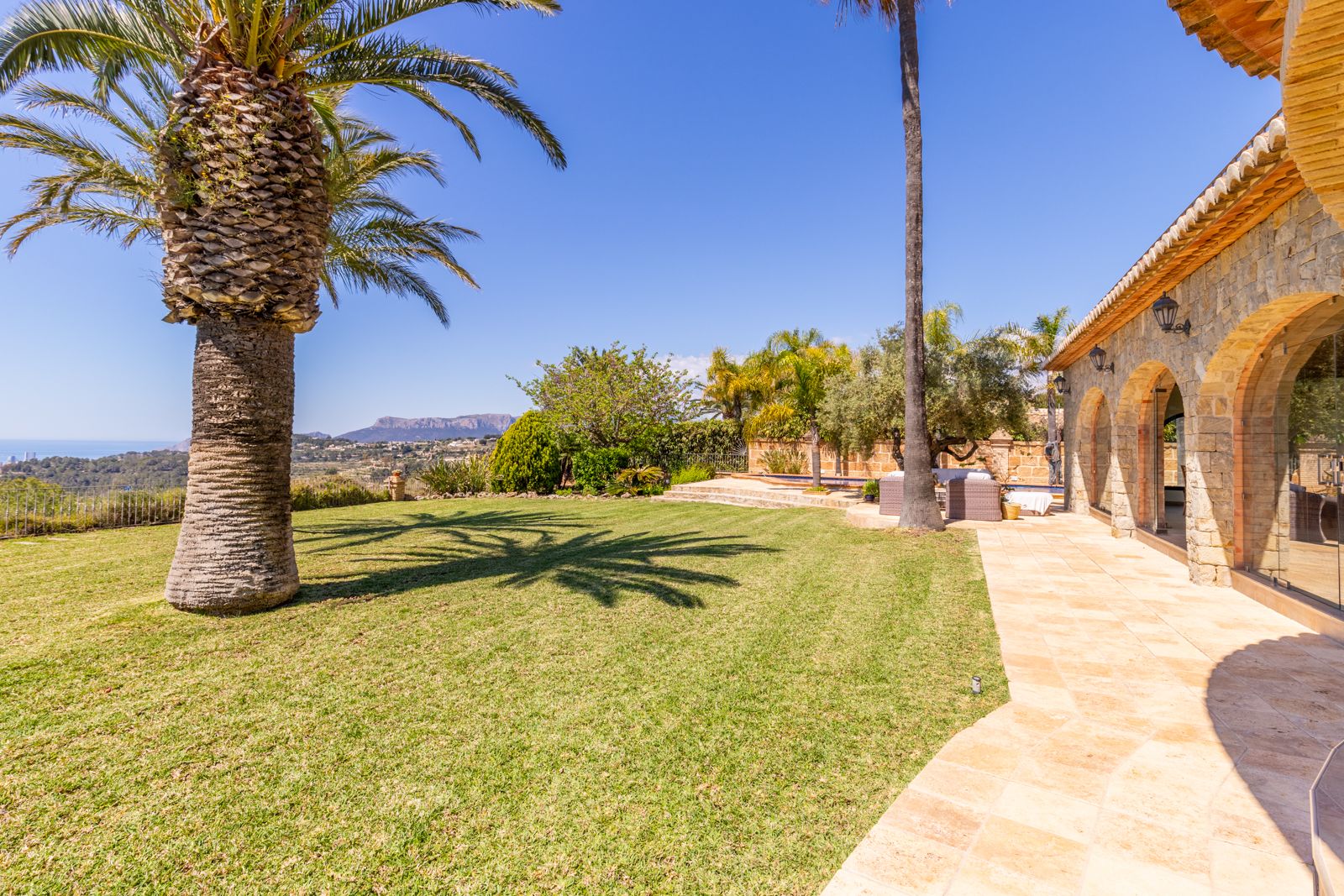 Vista del jardín con césped, palmeras y fachada de piedra con arcos.
