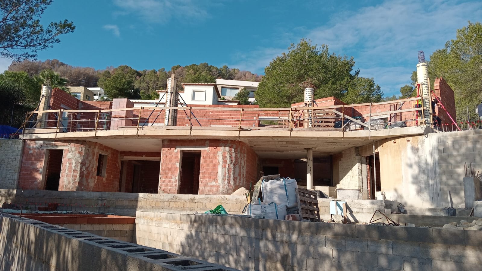 Vista de la estructura en construcción que muestra la planta inferior de ladrillo y la terraza superior curva, integrada en una parcela con muros de piedra.