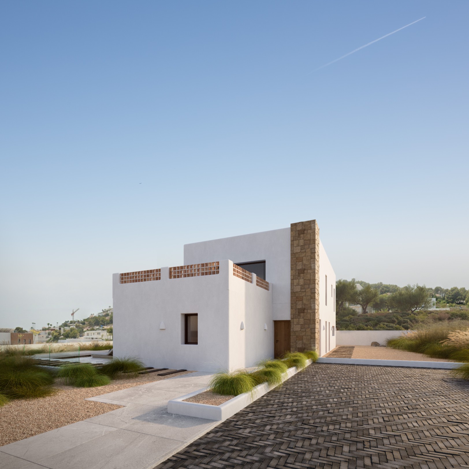 Fachada mediterránea moderna con muros blancos, un pilar de piedra natural y terraza con celosía de barro, rodeada de un jardín minimalista con pavimentos geométricos.