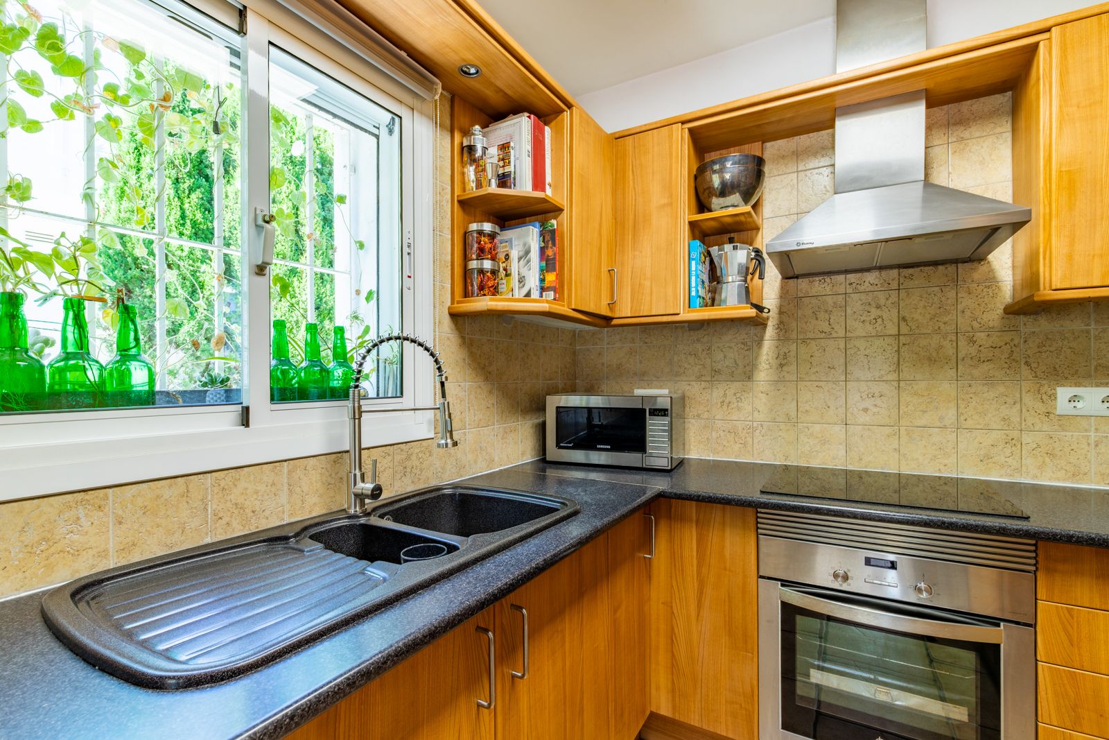 Cocina equipada con armarios de madera, encimera de granito oscuro y fregadero doble. Destaca la luz natural de la ventana y los electrodomésticos de acero inoxidable.