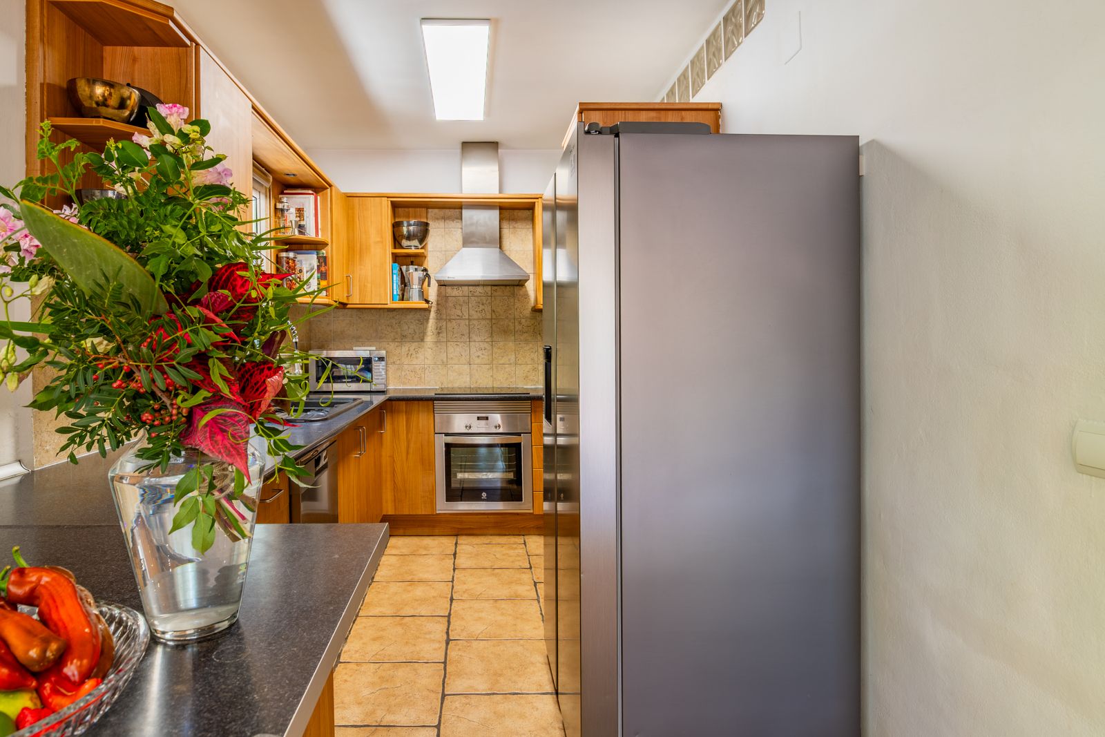 Cocina funcional con mobiliario de madera, encimera oscura y electrodomésticos de acero inoxidable. Suelo cerámico en tonos tierra y campana extractora decorativa.