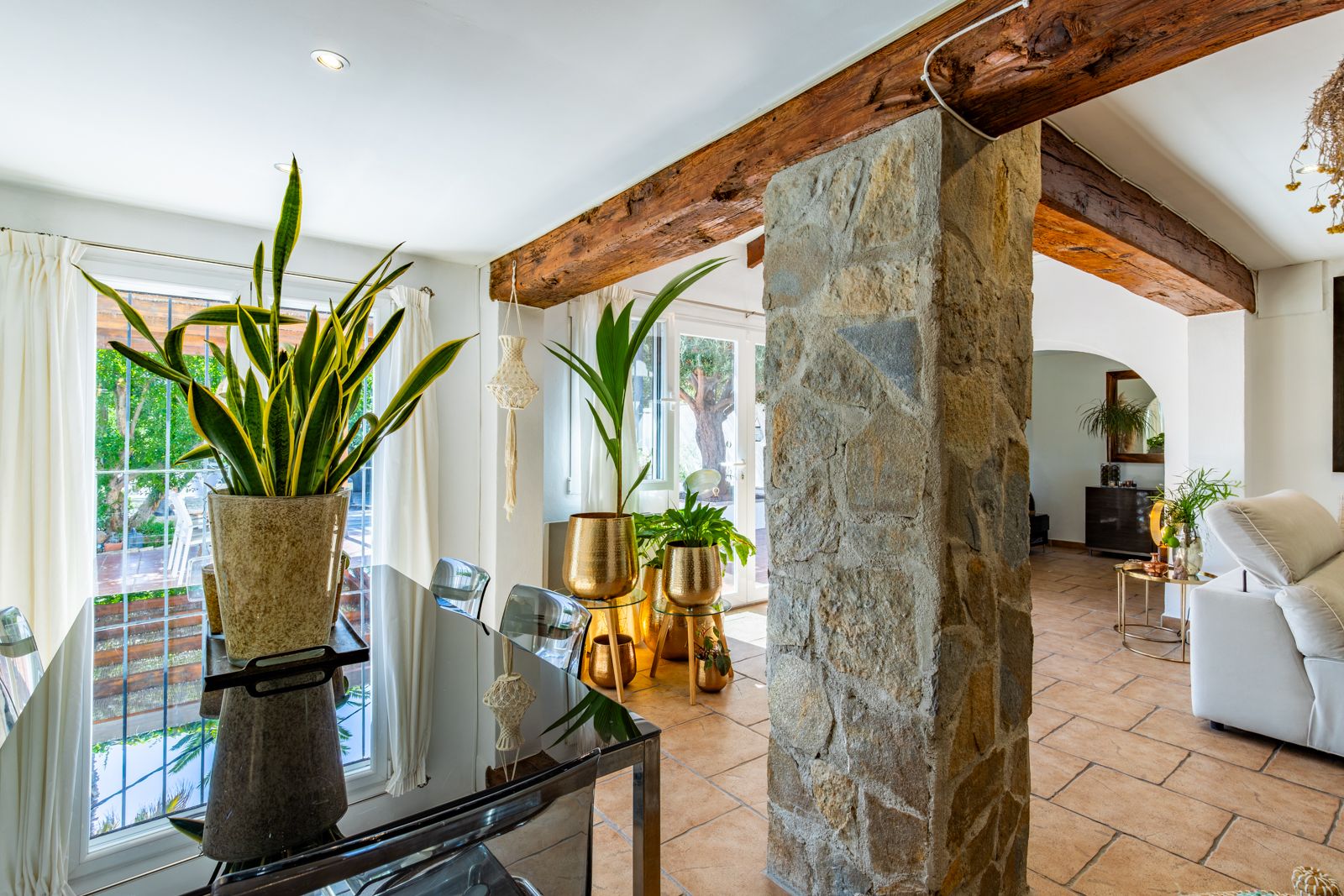Comedor con vigas de madera vista y pilar de piedra natural. Suelos cerámicos, mesa de cristal y grandes ventanales que integran el interior con el jardín.