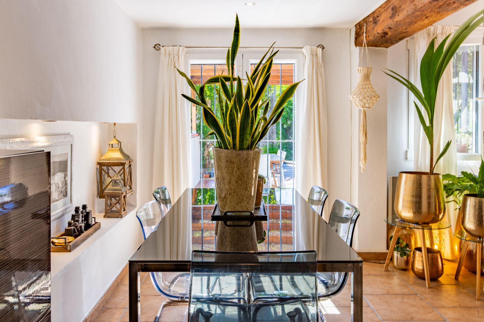 Comedor luminoso con mesa de cristal, sillas de policarbonato y vigas de madera vista. Grandes ventanales conectan el interior con la terraza y el jardín exterior.