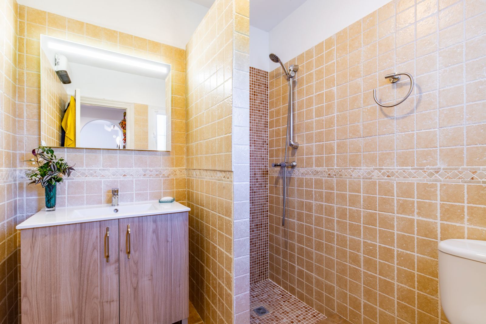 Cuarto de baño con ducha de obra revestida en gresite y azulejos en tonos tierra. Incluye mueble de lavabo con acabado en madera y espejo con iluminación integrada.