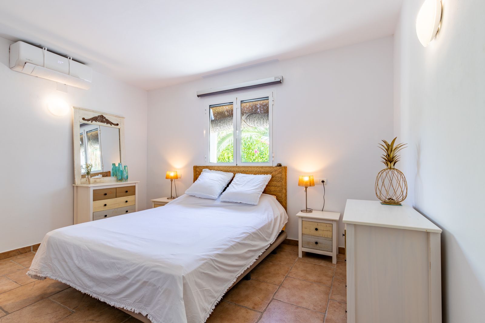 Dormitorio luminoso con suelos de terracota, aire acondicionado y mobiliario de estilo rústico. Cuenta con un cabecero de fibras naturales y ventana con vistas al jardín exterior.