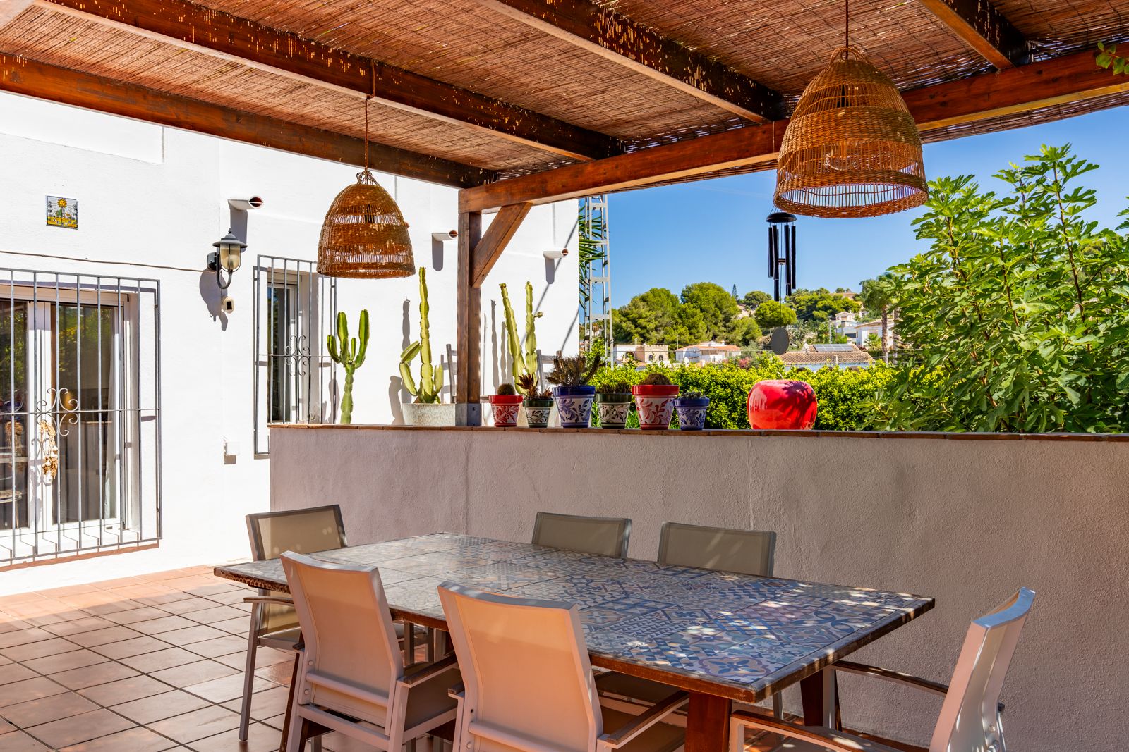 Terraza cubierta con pérgola de madera y cañizo, mesa de comedor con azulejos decorativos y lámparas de mimbre, con vistas al entorno natural de Baladrar.
