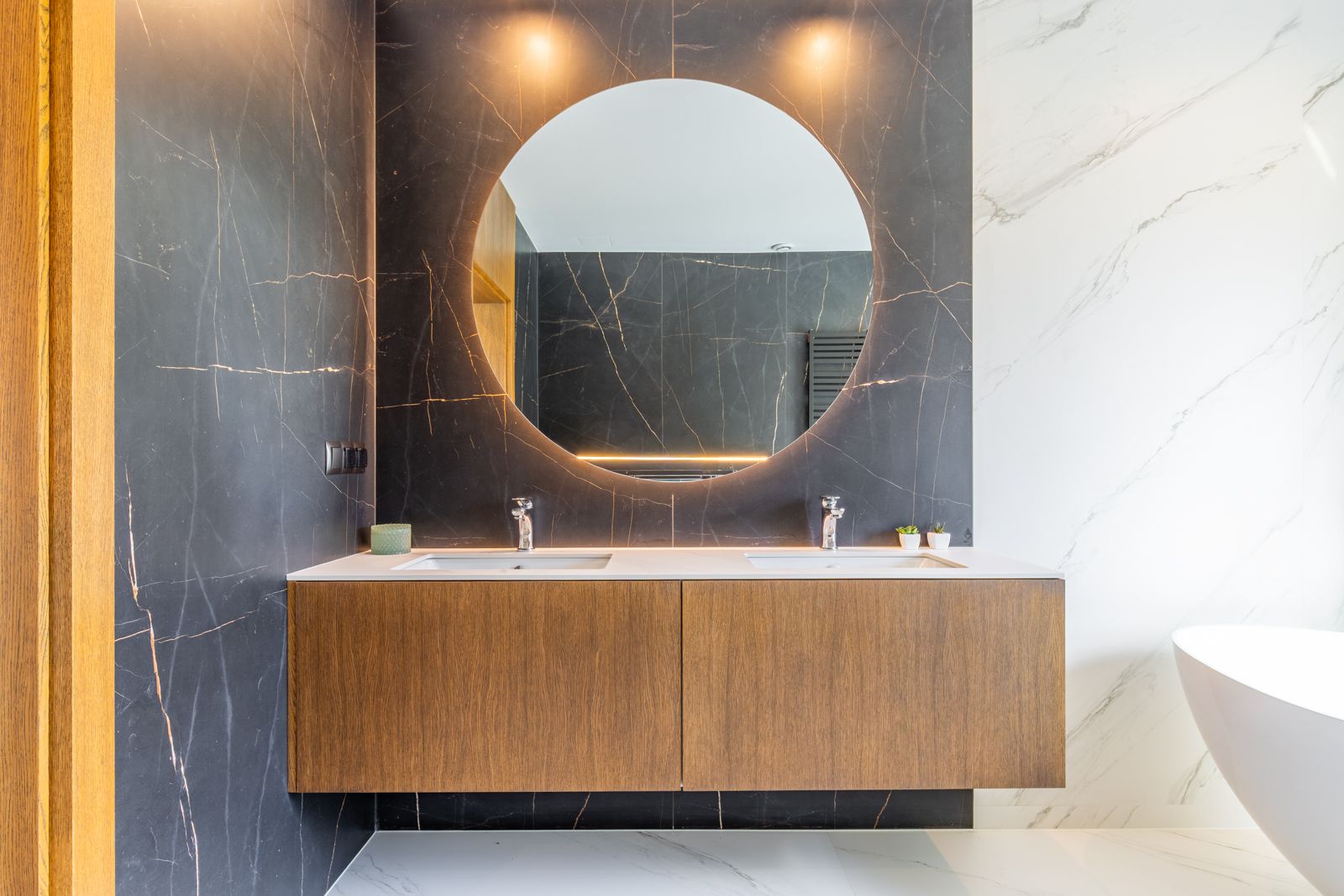 Baño de diseño con mueble de roble suspendido, lavabo de doble seno y espejo circular retroiluminado sobre revestimiento porcelánico oscuro con veteado dorado.