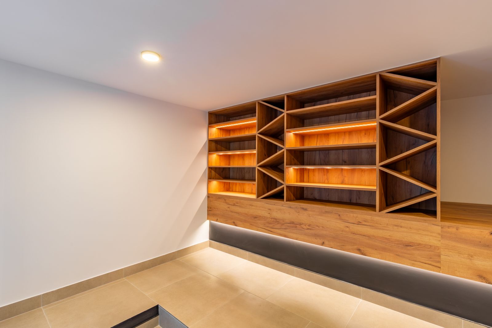 Bodega de diseño con estanterías de roble a medida, iluminación LED integrada y compartimentos geométricos sobre suelo de gres porcelánico.