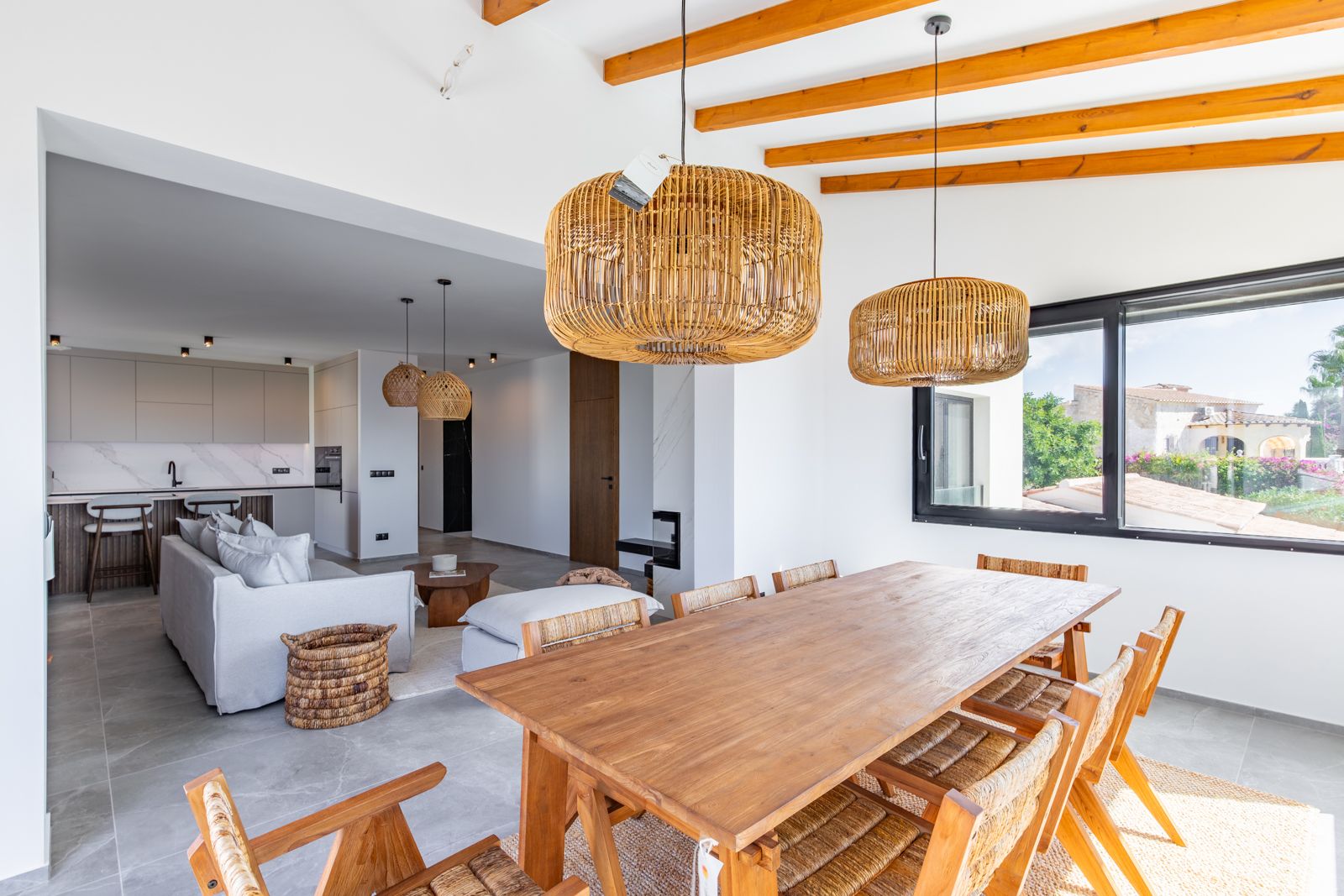 Comedor de concepto abierto con mesa de madera maciza, vigas vistas y lámparas de ratán, integrado con un salón minimalista y cocina de diseño.