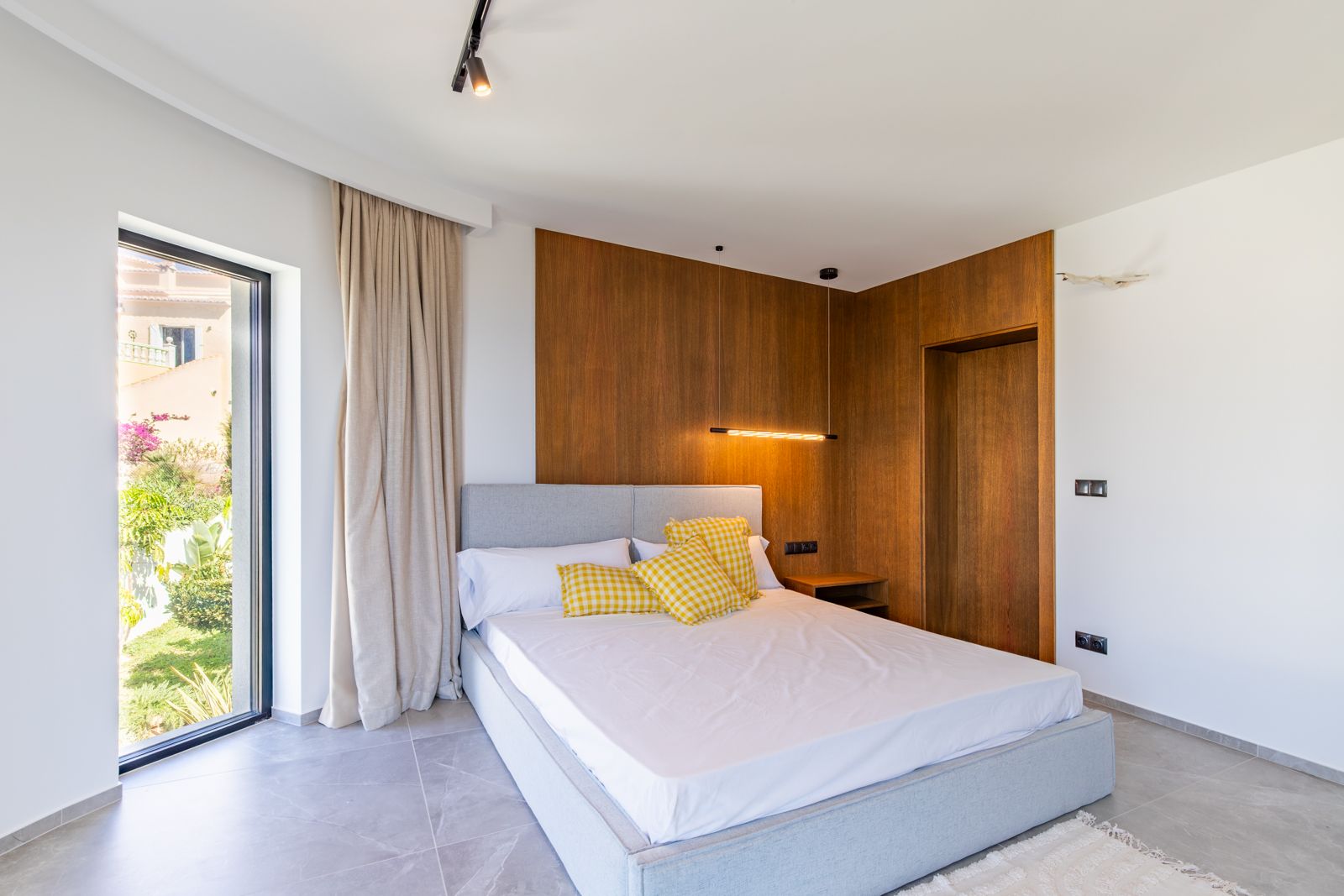 Dormitorio con panelado de roble natural, iluminación de diseño y acceso al jardín. Suelos de gres porcelánico de gran formato y ventanal técnico con vistas al exterior.