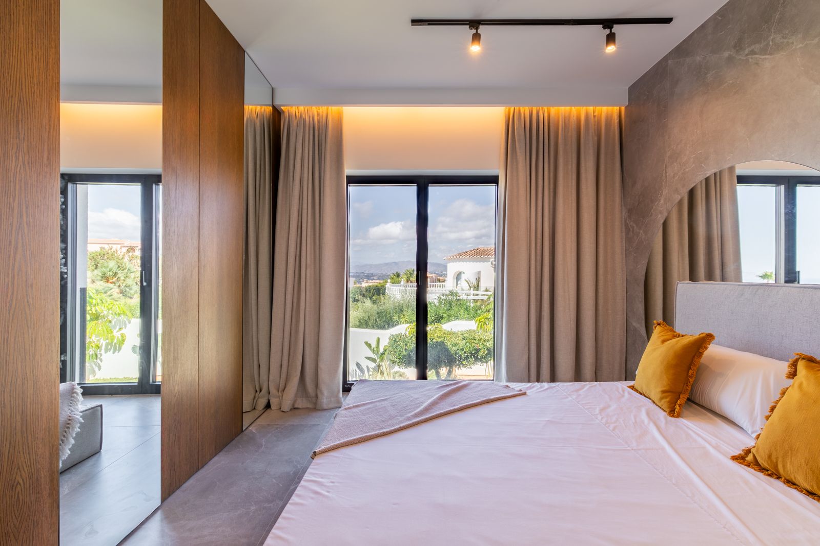 Dormitorio con paneles de roble natural, suelos de gres porcelánico y ventanales de gran formato con vistas al jardín y al paisaje mediterráneo.