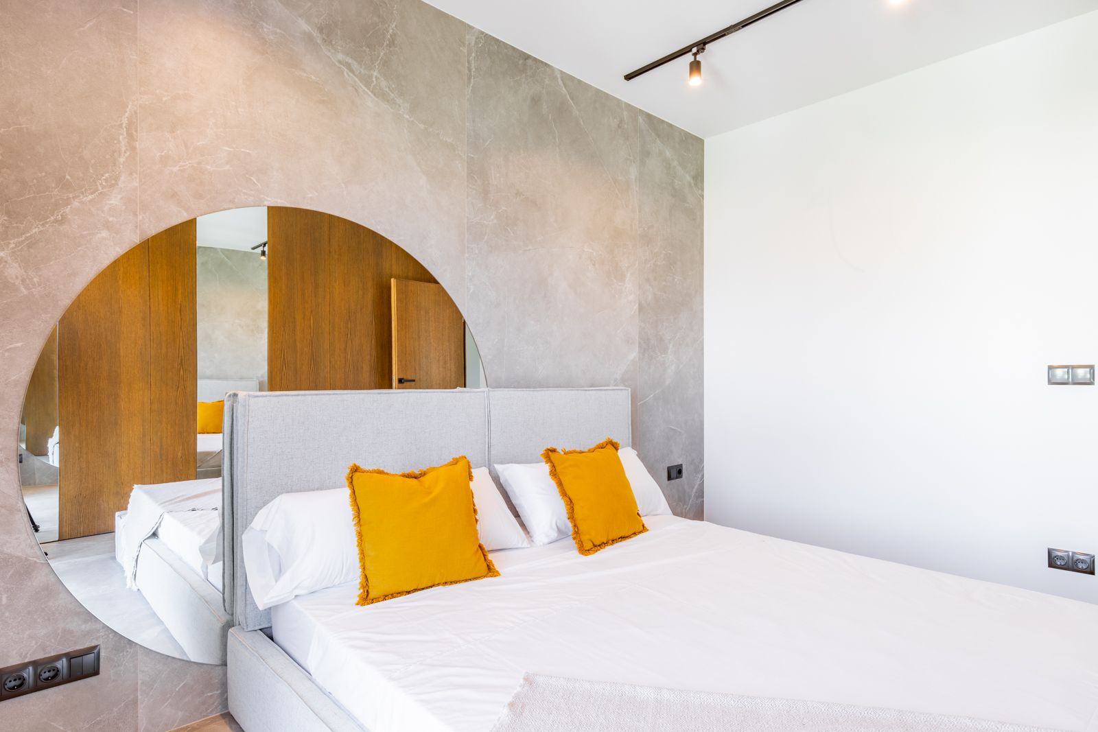 Dormitorio con pared de gres porcelánico de gran formato y espejo circular integrado. Incluye cabecero tapizado, paneles de roble natural y sistema de iluminación técnica.