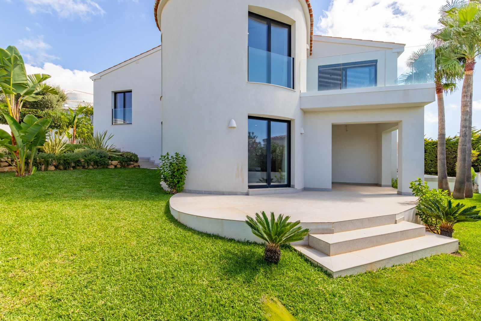 Fachada posterior con torre curva, carpintería de aluminio oscuro y terrazas con barandillas de cristal. El jardín cuenta con césped natural, palmeras y vegetación mediterránea.