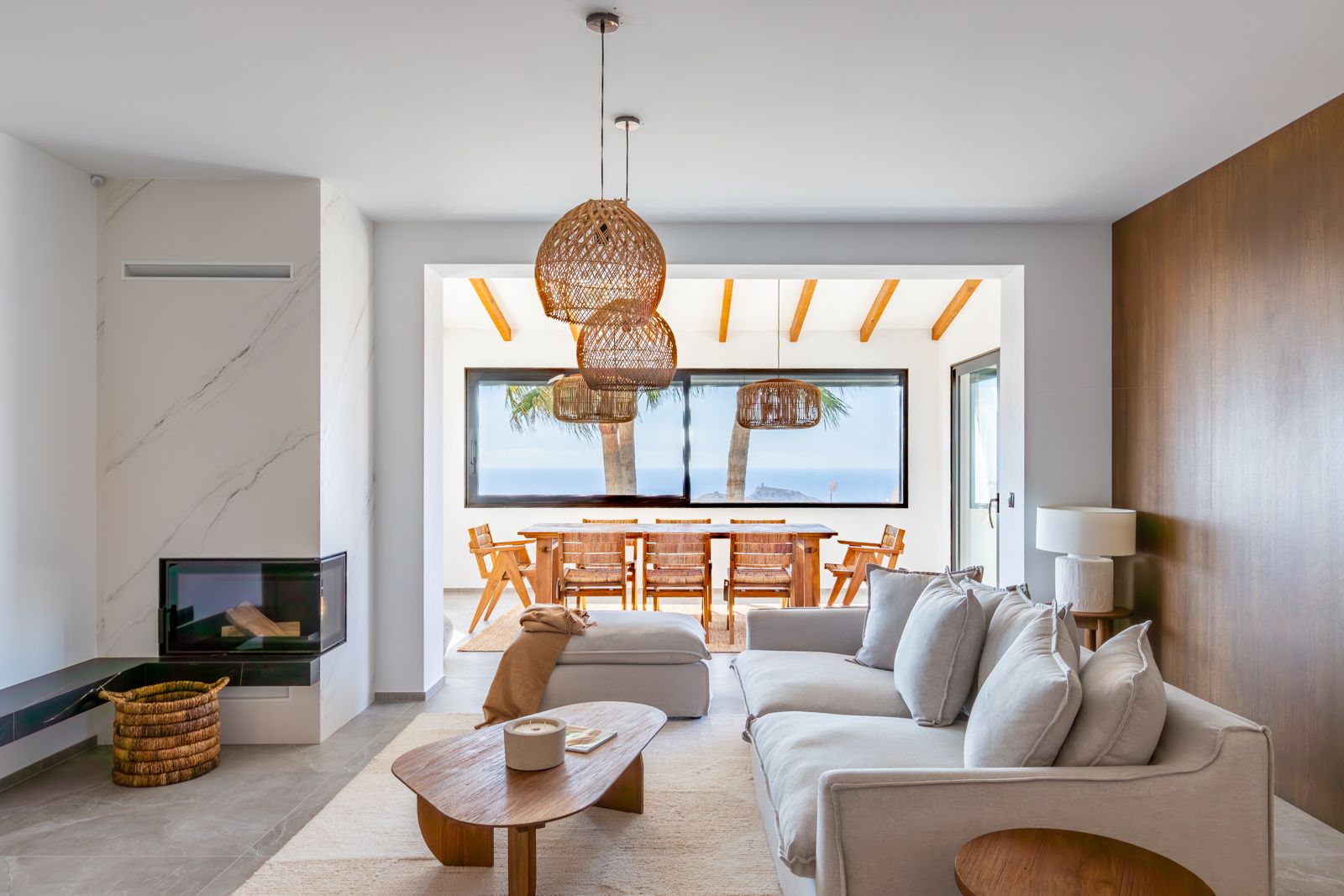 Salón con chimenea de piedra sinterizada, paneles de roble natural y zona de comedor con vistas al mar a través de grandes ventanales.