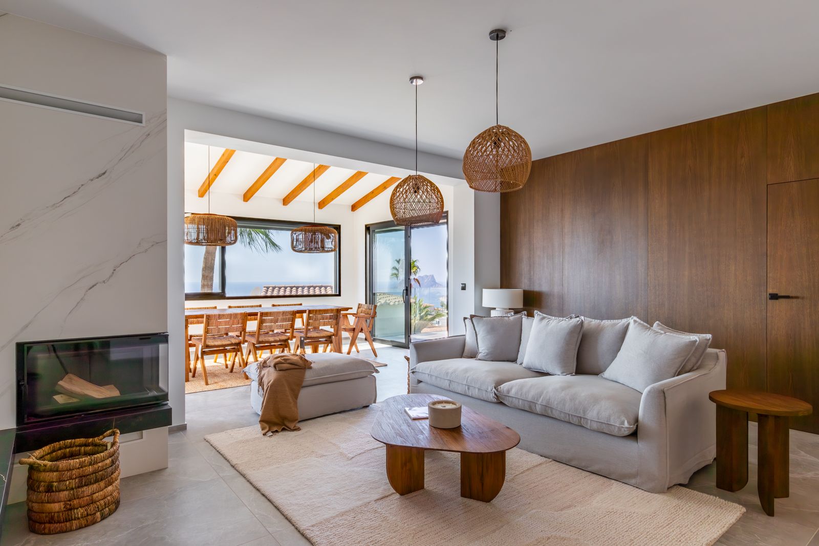 Salón con chimenea integrada, pared revestida en roble natural y vistas al mar. Destacan las lámparas de fibras naturales y el acceso al comedor con vigas de madera.