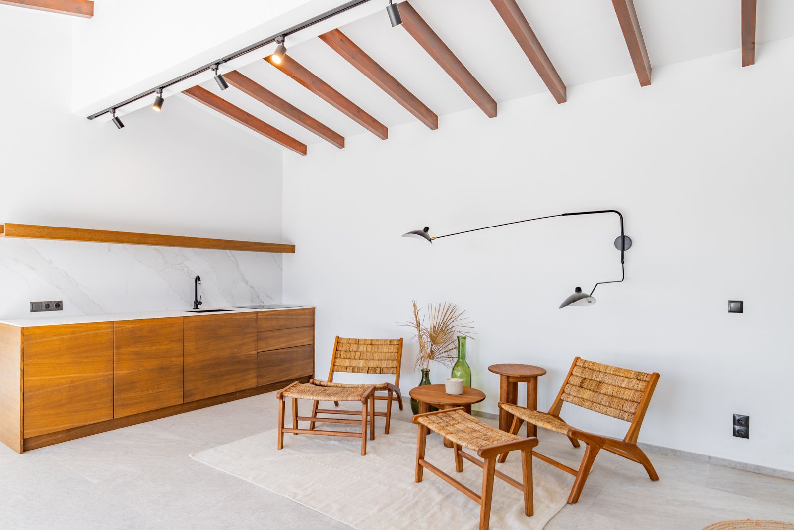 Salón con cocina integrada, vigas de madera vista, encimera de piedra sinterizada y mobiliario de roble con sillas de fibras naturales bajo iluminación técnica.
