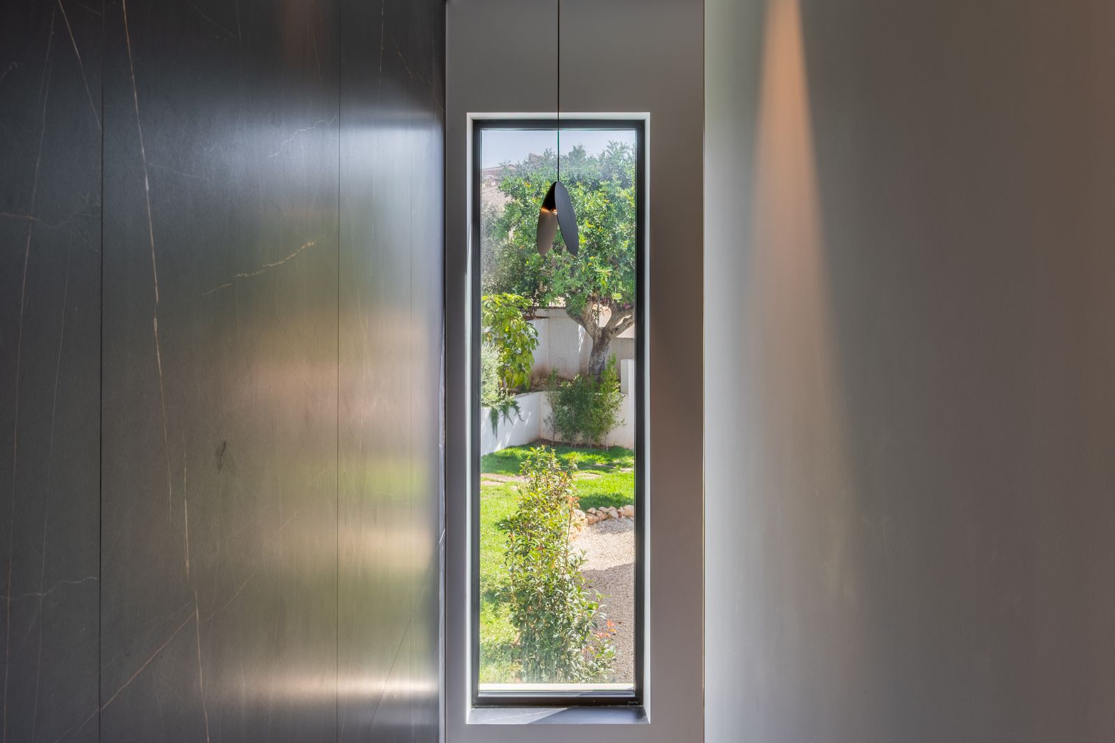 Ventanal vertical con triple acristalamiento que enmarca el jardín, junto a una pared de gres porcelánico oscuro de gran formato y luminaria de diseño minimalista.