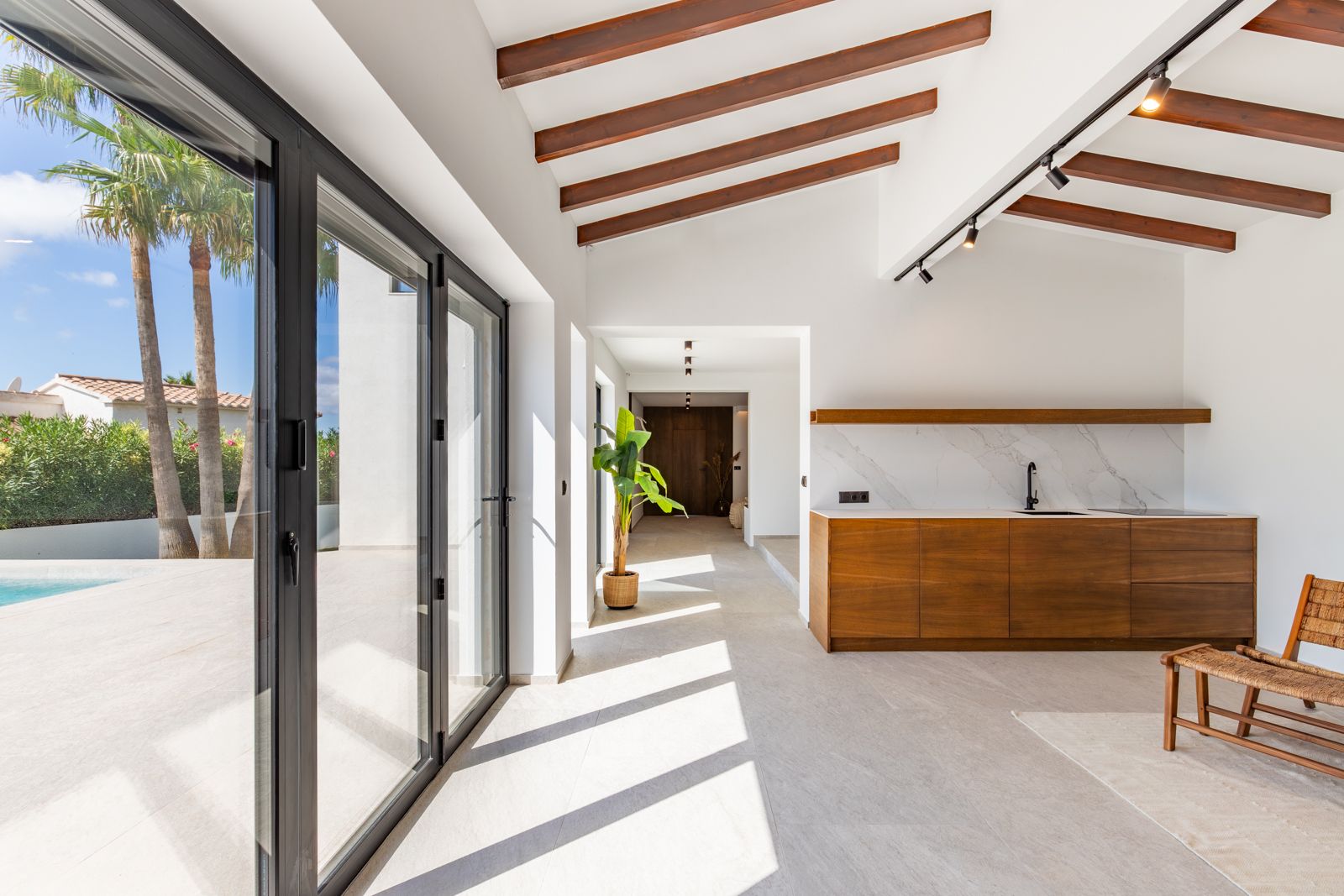 Zona de estar diáfana con vigas de madera, cocina integrada en roble con encimera de piedra y ventanales de gran formato con acceso directo a la terraza y piscina.