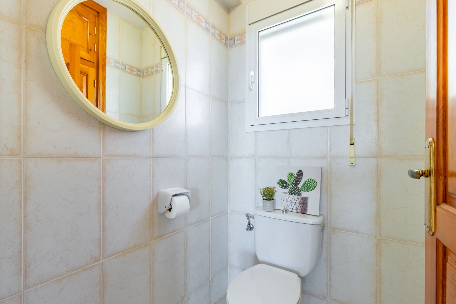 Aseo funcional con azulejos cerámicos en tonos beige, espejo circular de marco claro y ventana de ventilación que aporta luz natural al espacio.