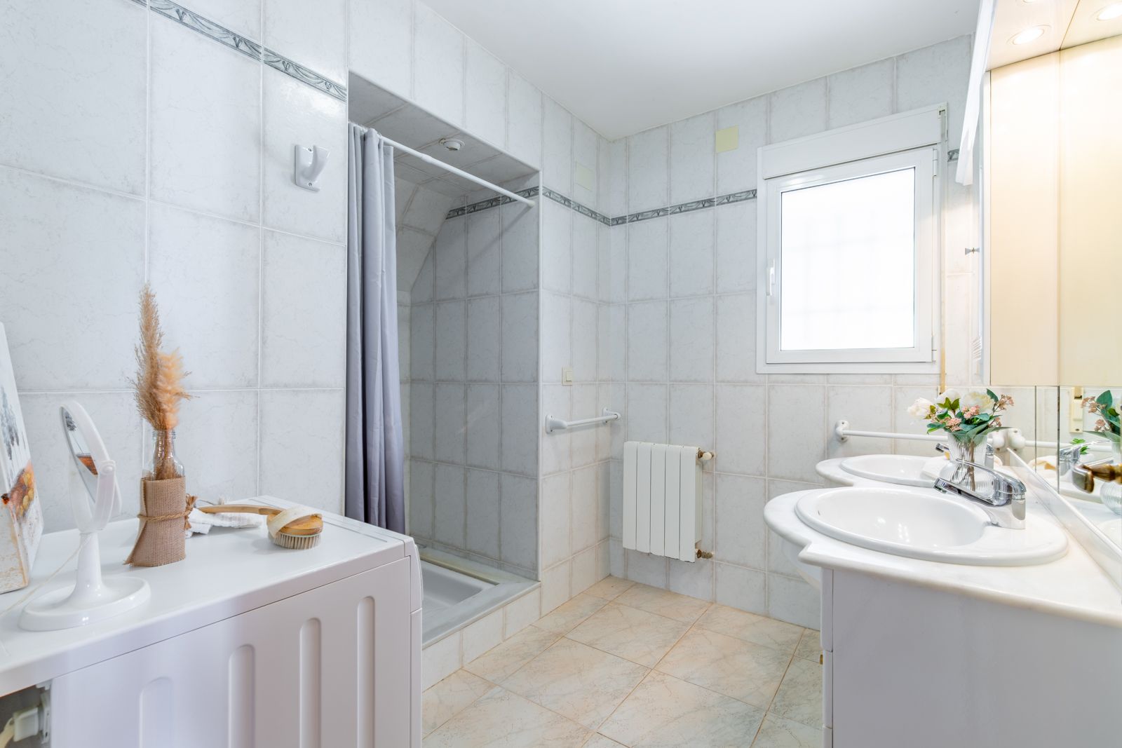 Baño funcional con encimera de mármol y doble seno, zona de lavandería integrada, ducha con cortina y radiador bajo la ventana de luz natural.
