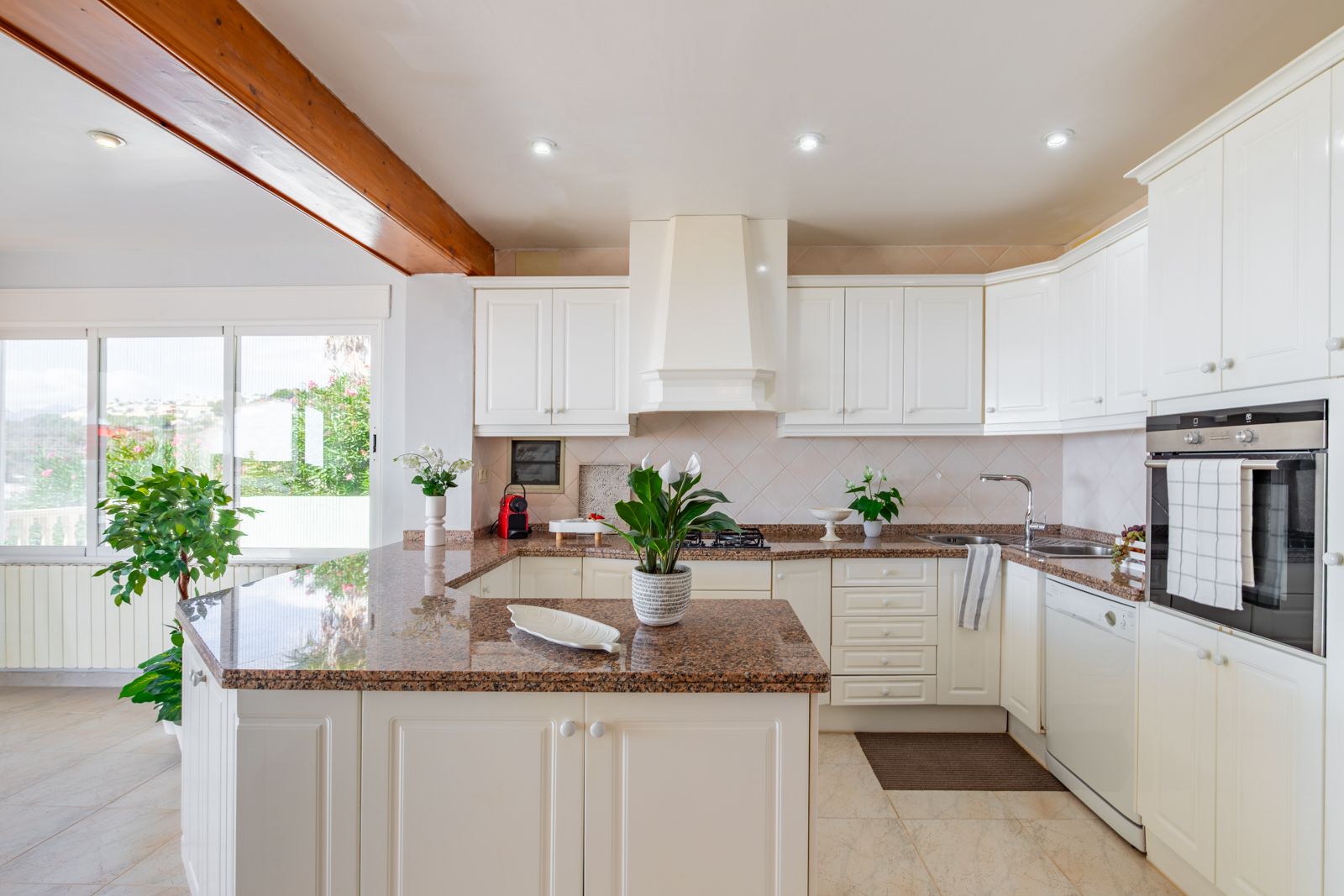 Cocina equipada con armarios blancos, encimera de granito y electrodomésticos integrados. Destaca su viga de madera vista, iluminación empotrada y acceso directo al exterior.