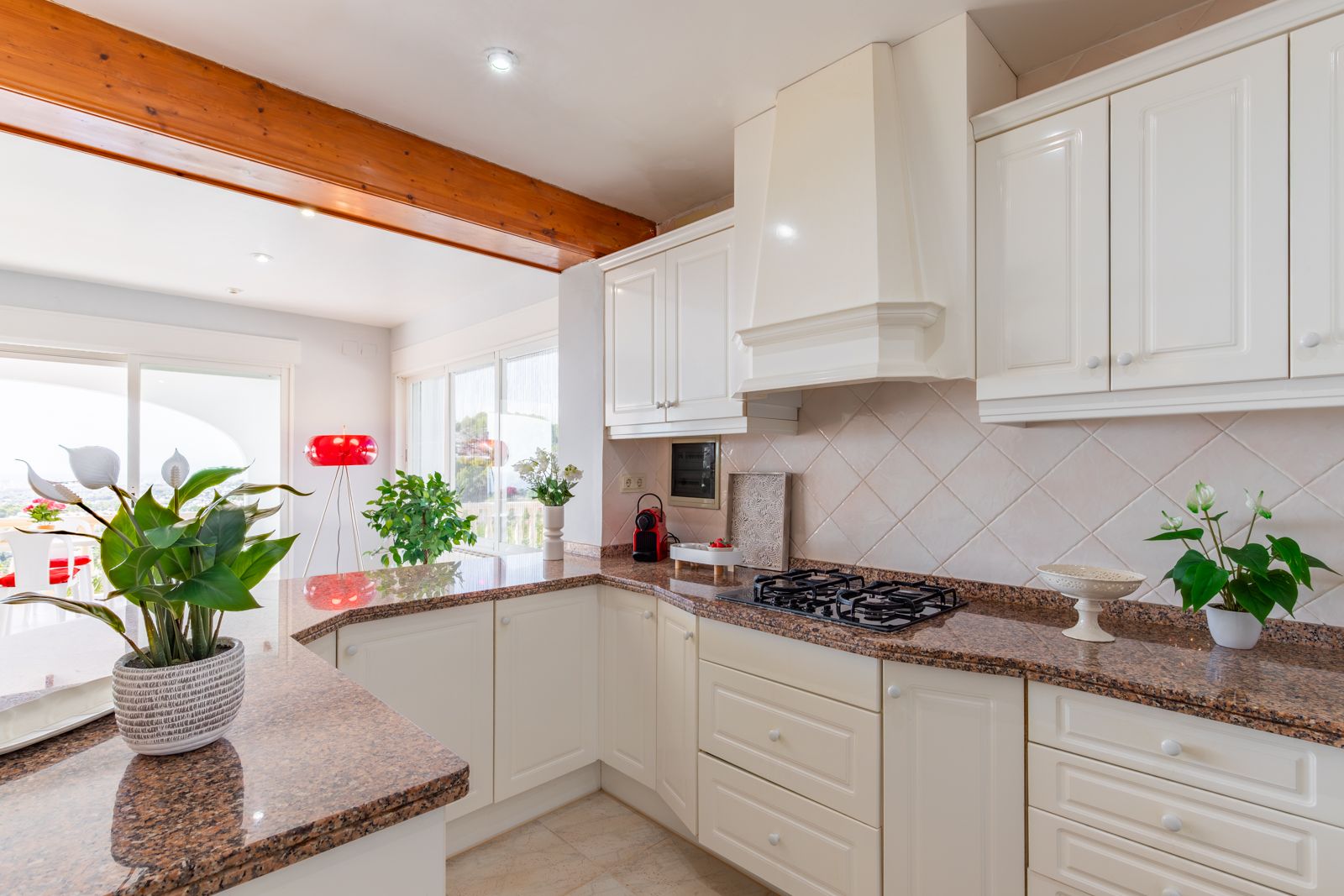 Cocina equipada con encimera de granito, armarios blancos de estilo clásico y vigas de madera vistas, abierta hacia un luminoso salón comedor.