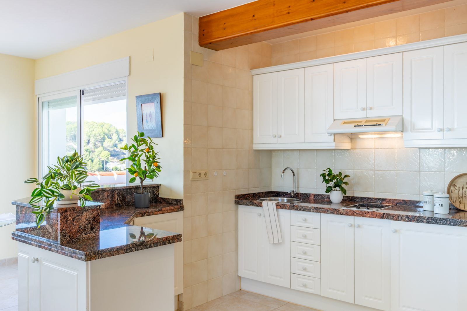 Cocina funcional con encimeras de granito oscuro, armarios blancos y vigas de madera vistas. Dispone de luz natural abundante y vistas despejadas a través del ventanal.